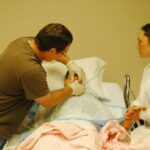 A nursing student in a white coat observes as an instructor demonstrates patient care techniques on a hospital bed with medical equipment nearby.