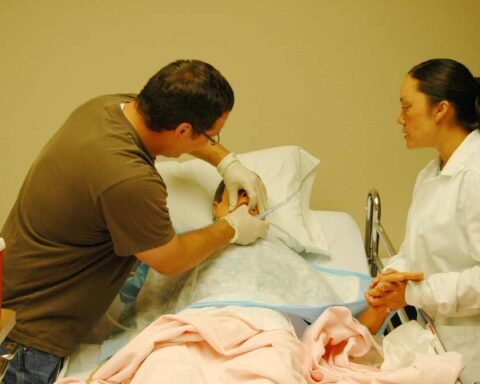 A nursing student in a white coat observes as an instructor demonstrates patient care techniques on a hospital bed with medical equipment nearby.