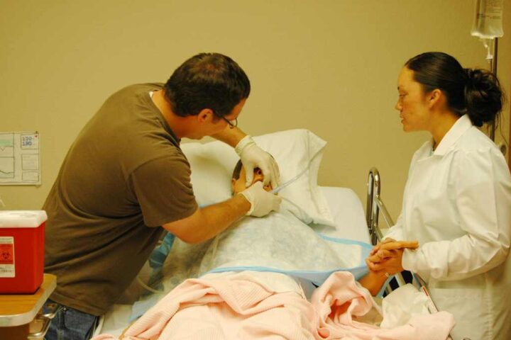 A nursing student in a white coat observes as an instructor demonstrates patient care techniques on a hospital bed with medical equipment nearby.