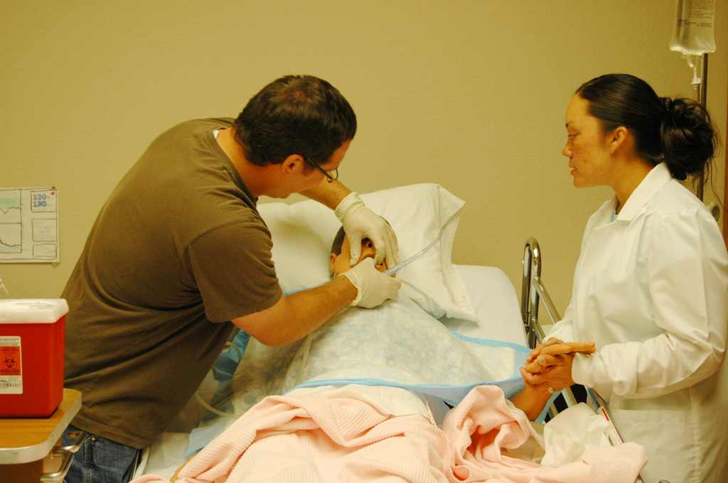 A nursing student in a white coat observes as an instructor demonstrates patient care techniques on a hospital bed with medical equipment nearby.