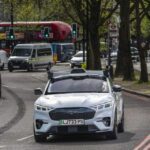 A white Ford Mustang Mach-E with autonomous driving sensors mounted on top drives on a London street with red buses visible in the background.