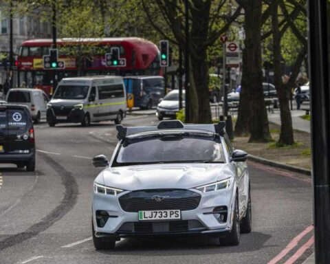A white Ford Mustang Mach-E with autonomous driving sensors mounted on top drives on a London street with red buses visible in the background.