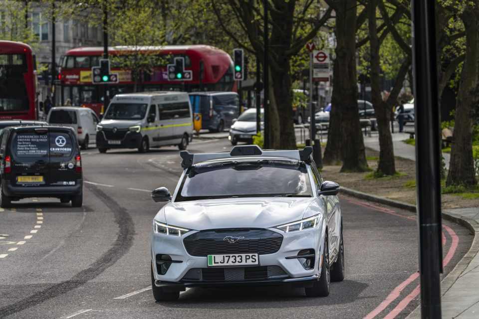 A white Ford Mustang Mach-E with autonomous driving sensors mounted on top drives on a London street with red buses visible in the background.