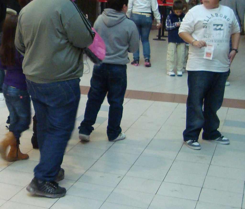Several people with varying body types standing in what appears to be a shopping mall or public space, photographed from behind.