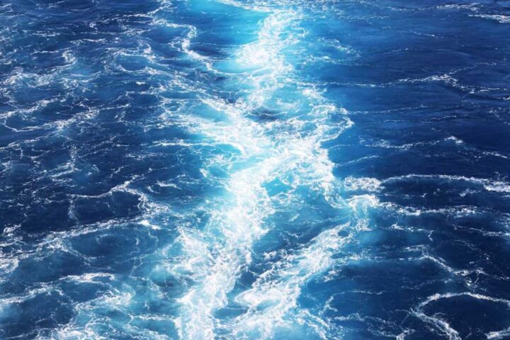 Aerial view of deep blue ocean water with white foam creating a pattern through the center, showing waves and currents on the open sea.