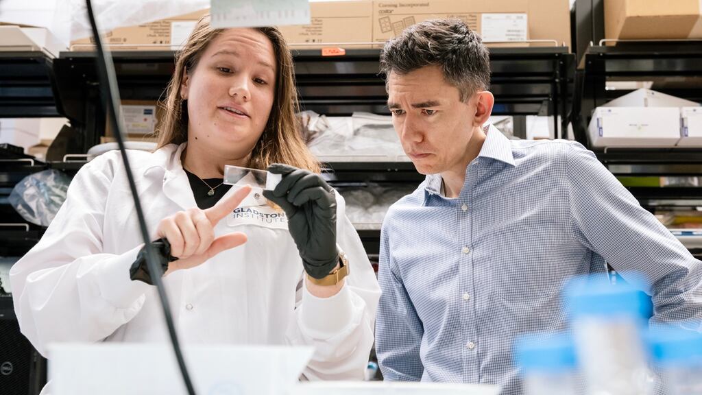 Two researchers in a laboratory setting - a female scientist in a white lab coat on the left and a male scientist in a blue checkered shirt on the right, examining laboratory equipment in what appears to be a neuroscience research facility