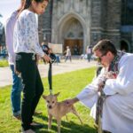 Clergy member in white robe blessing a small dog held on a leash by a woman outside Duke Chapel during the annual pet blessing event.