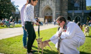 Clergy member in white robe blessing a small dog held on a leash by a woman outside Duke Chapel during the annual pet blessing event.