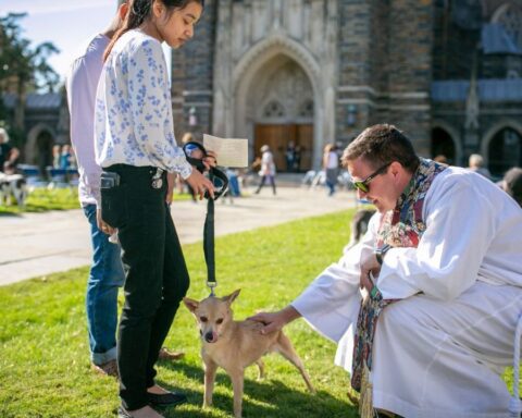 Clergy member in white robe blessing a small dog held on a leash by a woman outside Duke Chapel during the annual pet blessing event.