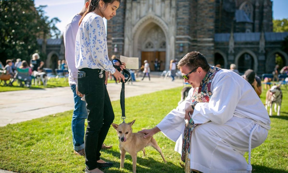 Clergy member in white robe blessing a small dog held on a leash by a woman outside Duke Chapel during the annual pet blessing event.