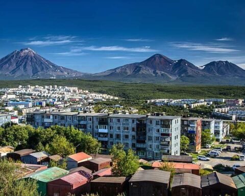 Panoramic view of Petropavlovsk-Kamchatsky city with apartment buildings in the foreground and the snow-capped volcanoes Avachinsky and Koryaksky dominating the background landscape.