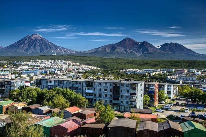 Panoramic view of Petropavlovsk-Kamchatsky city with apartment buildings in the foreground and the snow-capped volcanoes Avachinsky and Koryaksky dominating the background landscape.