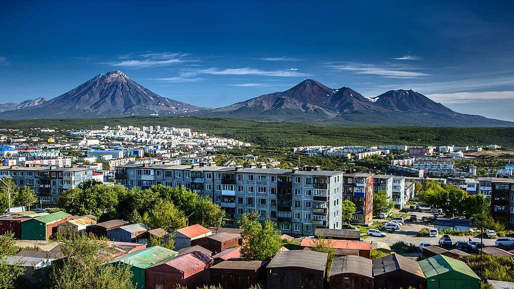 Panoramic view of Petropavlovsk-Kamchatsky city with apartment buildings in the foreground and the snow-capped volcanoes Avachinsky and Koryaksky dominating the background landscape.