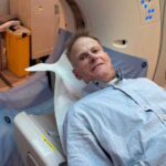 Professor Richard Scolyer lying on a Canon Aquilion CT scanner bed in a hospital room, wearing a light blue hospital gown, preparing for a brain scan as part of his glioblastoma treatment.