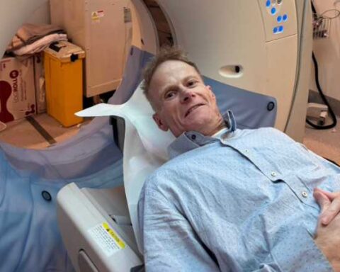 Professor Richard Scolyer lying on a Canon Aquilion CT scanner bed in a hospital room, wearing a light blue hospital gown, preparing for a brain scan as part of his glioblastoma treatment.