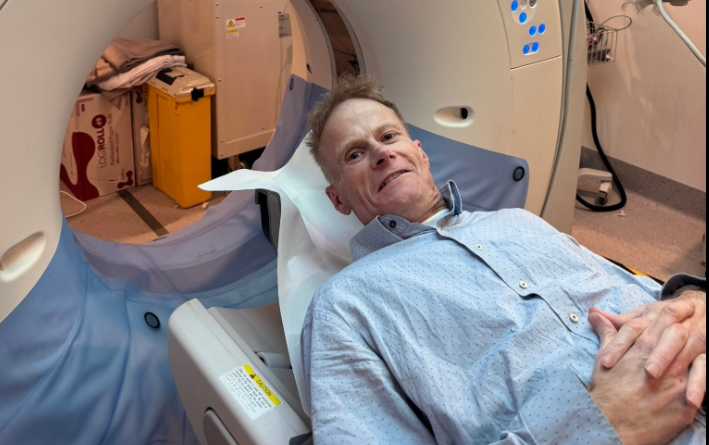 Professor Richard Scolyer lying on a Canon Aquilion CT scanner bed in a hospital room, wearing a light blue hospital gown, preparing for a brain scan as part of his glioblastoma treatment.