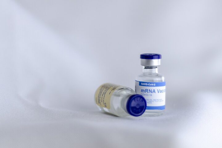 Two vaccine vials with blue caps, one standing upright and one lying on its side, labeled as mRNA vaccine against a light blue background.