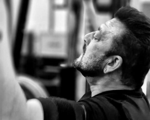 Black-and-white photo of Sanjay Dutt working out in a gym, eyes closed in focus, lifting weights with intensity.
