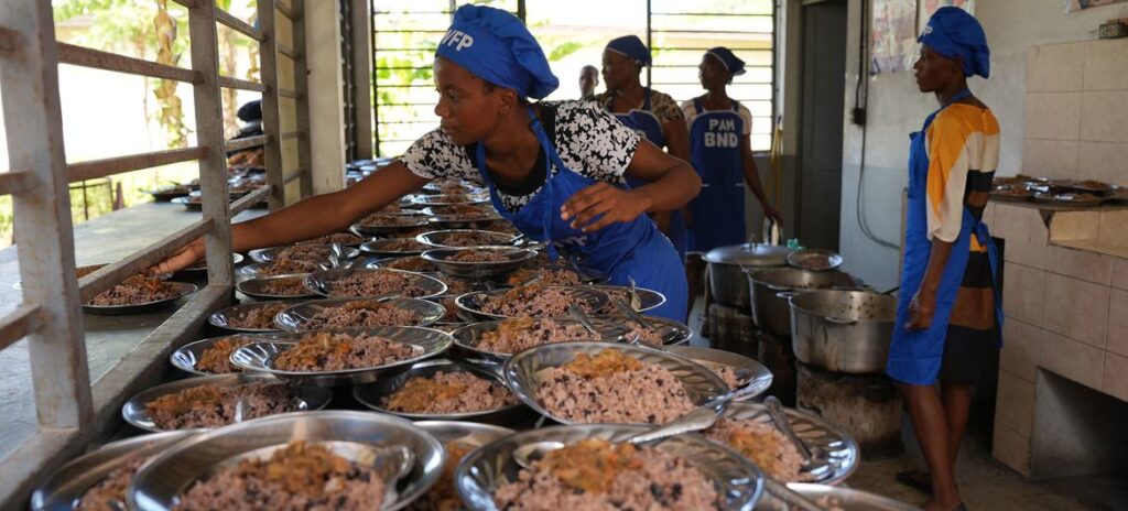 Female cooks in blue WFP uniforms preparing and serving rice meals for students in a school kitchen in Haiti, with multiple metal plates of food lined up on a serving counter.