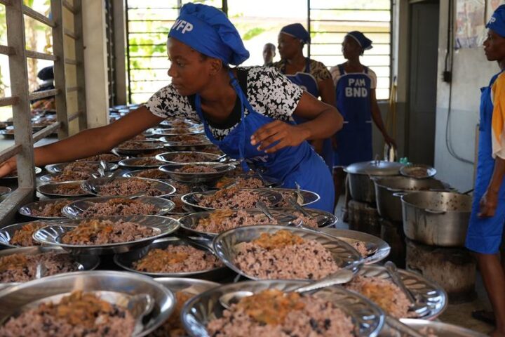 Female cooks in blue WFP uniforms preparing and serving rice meals for students in a school kitchen in Haiti, with multiple metal plates of food lined up on a serving counter.