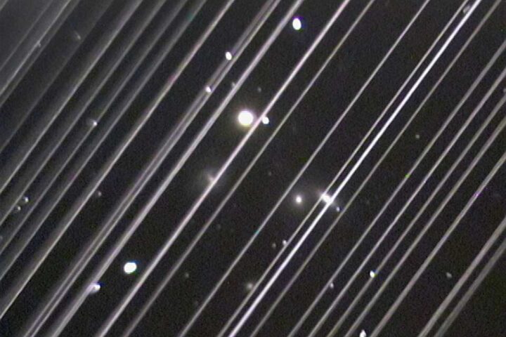 Long‑exposure photograph of the night sky showing diagonal bright trails from passing Starlink satellites against a backdrop of stars and the NGC 5353/4 galaxy group.