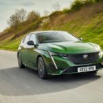Green Peugeot 308 hatchback driving on a highway with grassy hillside in background