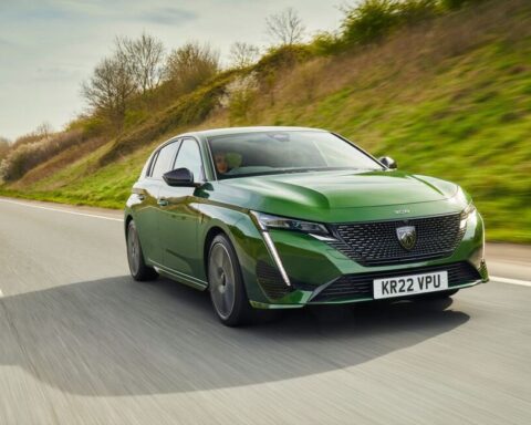 Green Peugeot 308 hatchback driving on a highway with grassy hillside in background