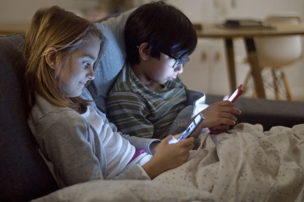 Two pre-teen children, a girl with light brown hair and a boy with dark hair and glasses, sitting close together under a blanket on a couch, each looking at their own mobile devices with screens illuminating their faces.