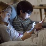 Two pre-teen children, a girl with light brown hair and a boy with dark hair and glasses, sitting close together under a blanket on a couch, each looking at their own mobile devices with screens illuminating their faces.