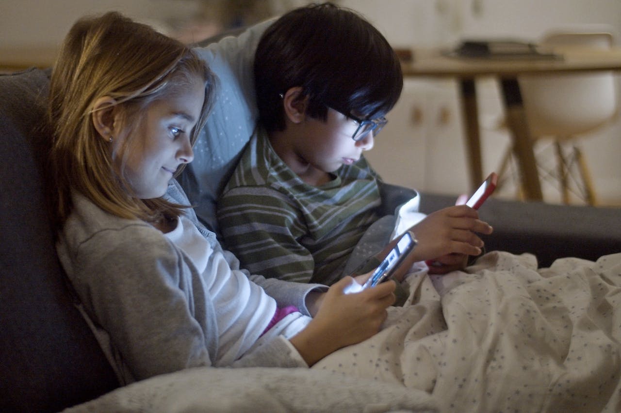 Two pre-teen children, a girl with light brown hair and a boy with dark hair and glasses, sitting close together under a blanket on a couch, each looking at their own mobile devices with screens illuminating their faces.
