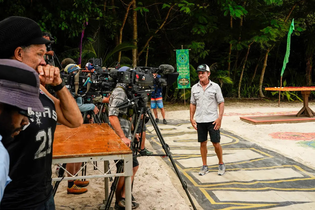 Survivor production crew filming a host on a tribal-themed set in Fiji’s Mamanuca Islands, with cameras, banners, and tropical backdrop.