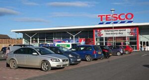 Exterior of a Tesco supermarket with red logo signage and cars parked in the foreground.