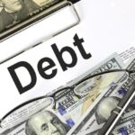 Close-up image of a document with the word "Debt" prominently displayed, overlaid with US dollar bills, reading glasses, and a pen, representing financial obligations and paperwork associated with debt management.