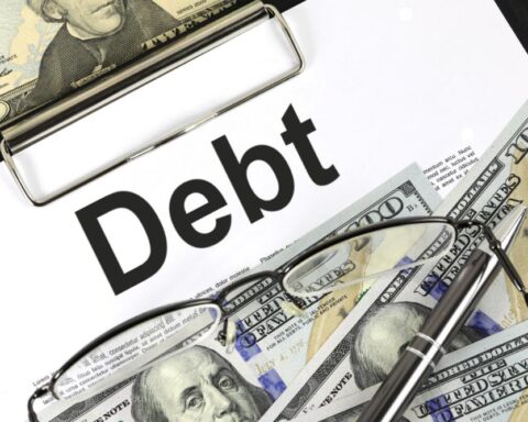 Close-up image of a document with the word "Debt" prominently displayed, overlaid with US dollar bills, reading glasses, and a pen, representing financial obligations and paperwork associated with debt management.