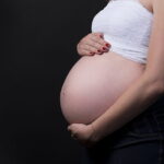 Side profile of a pregnant woman in a white crop top holding her belly against a black background