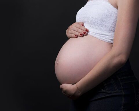 Side profile of a pregnant woman in a white crop top holding her belly against a black background