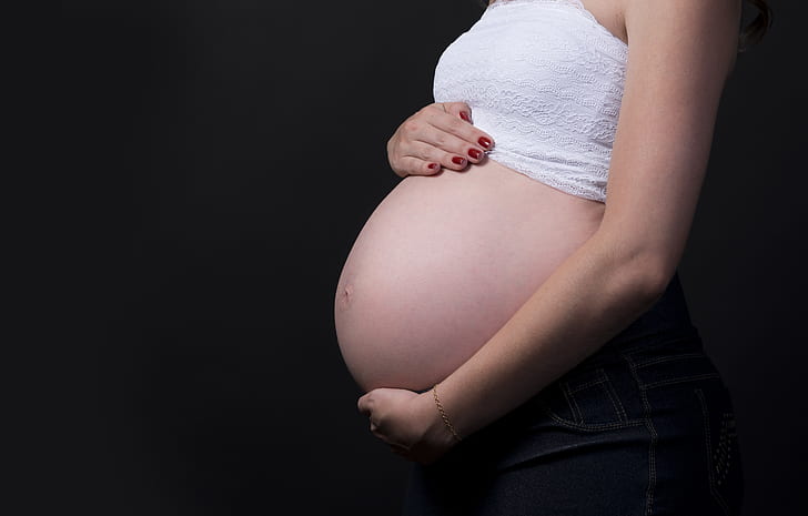 Side profile of a pregnant woman in a white crop top holding her belly against a black background