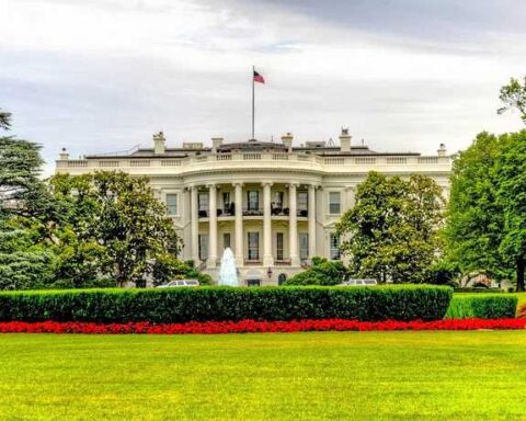 The White House south facade with its iconic white columns and portico, surrounded by green lawns, trees, and red flowers in the foreground.