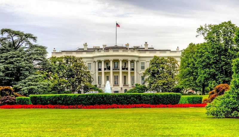 The White House south facade with its iconic white columns and portico, surrounded by green lawns, trees, and red flowers in the foreground.