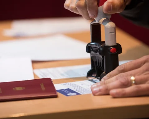 Hand using an official stamp on a passport with travel documents visible on a wooden desk.