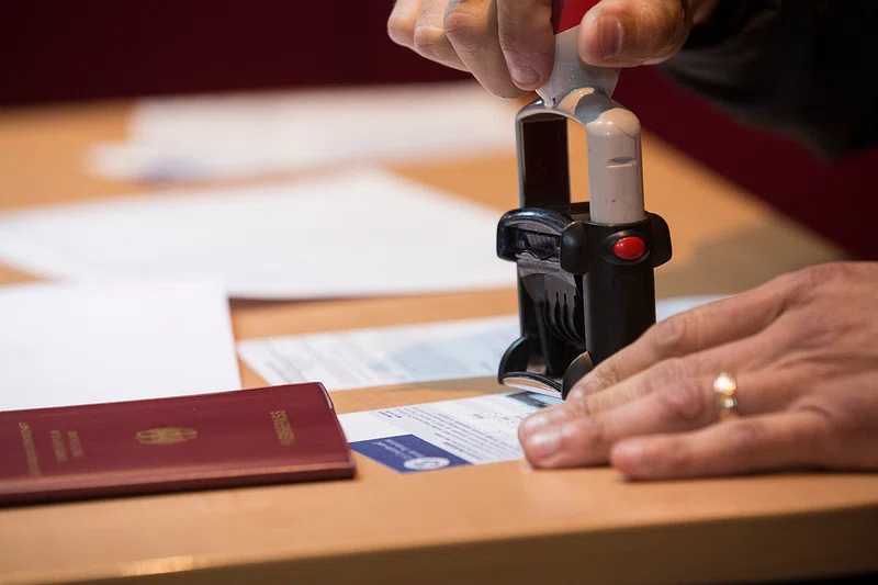 Hand using an official stamp on a passport with travel documents visible on a wooden desk.