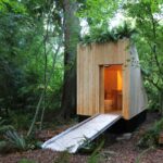 Wooden composting toilet structure with wheelchair-accessible ramp nestled among trees in UBC Botanical Garden, featuring a green roof with plants growing on top.