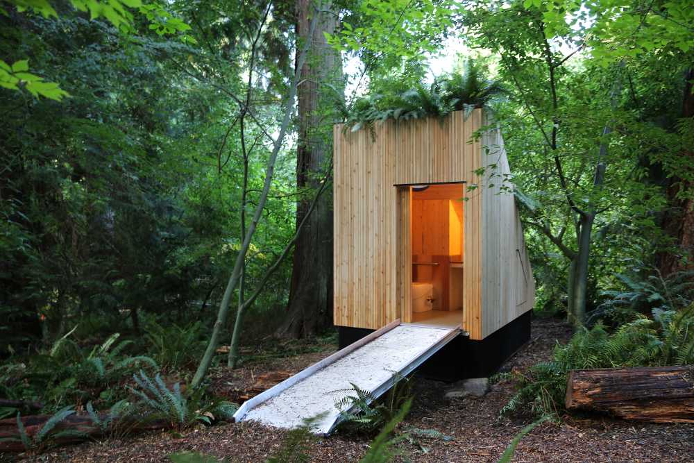 Wooden composting toilet structure with wheelchair-accessible ramp nestled among trees in UBC Botanical Garden, featuring a green roof with plants growing on top.