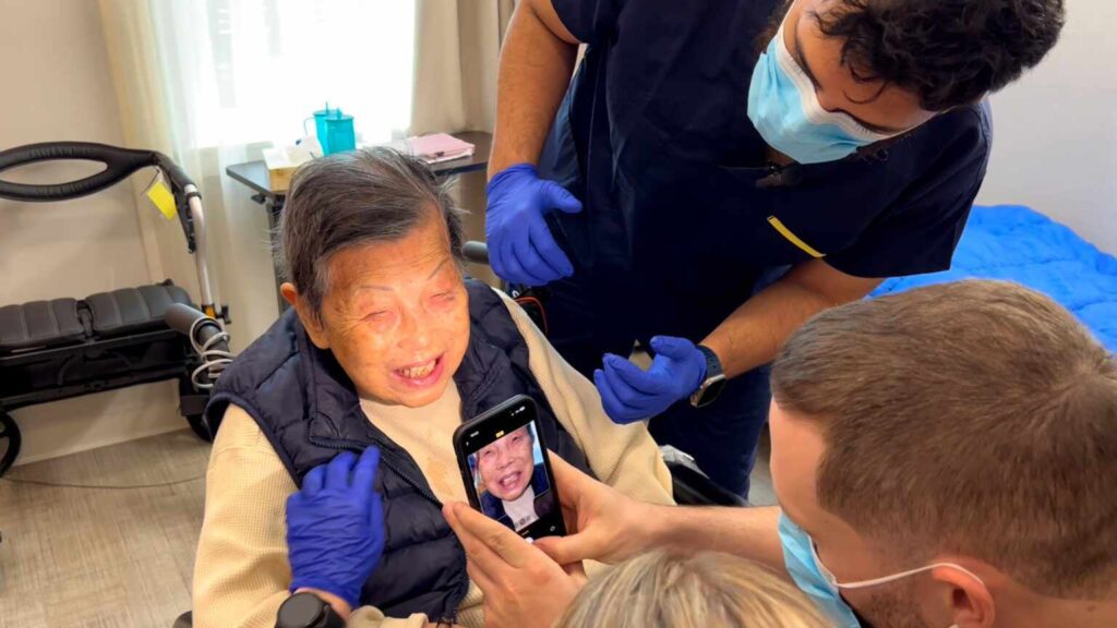 Dental students at UBC capture digital images of an elderly patient's teeth using a smartphone during an examination at Villa Cathay Care Home. One student holds a smartphone displaying the patient's face while another examines her teeth.