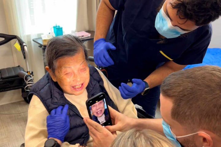 Dental students at UBC capture digital images of an elderly patient's teeth using a smartphone during an examination at Villa Cathay Care Home. One student holds a smartphone displaying the patient's face while another examines her teeth.