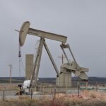 An oil pumpjack (nodding donkey) operating in an arid landscape with sparse vegetation and utility poles in the background under an overcast sky.
