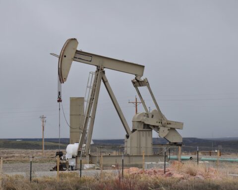 An oil pumpjack (nodding donkey) operating in an arid landscape with sparse vegetation and utility poles in the background under an overcast sky.