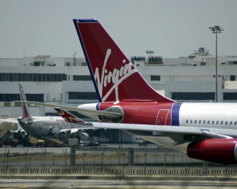 Virgin Australia aircraft tail featuring the red and white Virgin logo, parked at an airport with terminal buildings visible in the background.
