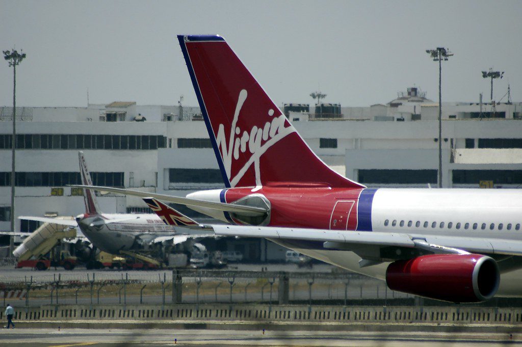 Virgin Australia aircraft tail featuring the red and white Virgin logo, parked at an airport with terminal buildings visible in the background.