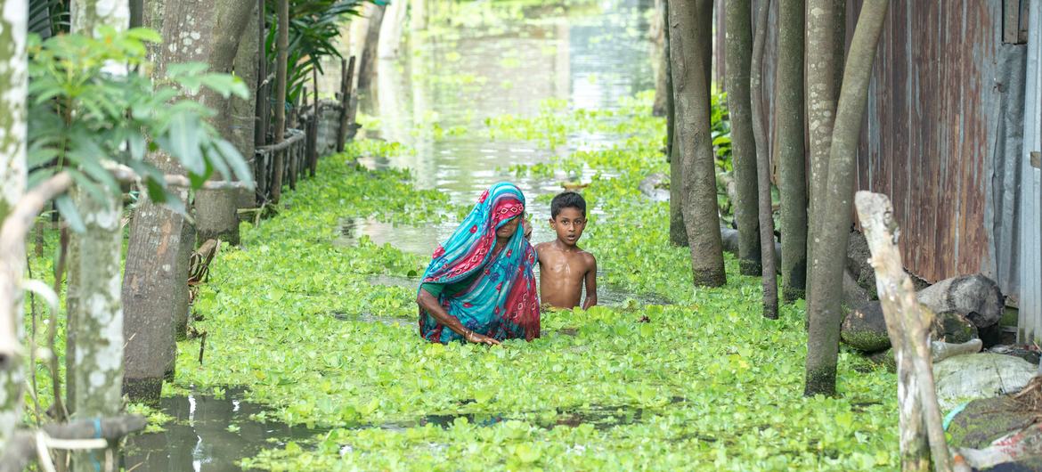 A woman and child wade through flood waters filled with green vegetation in Kurigram, Bangladesh